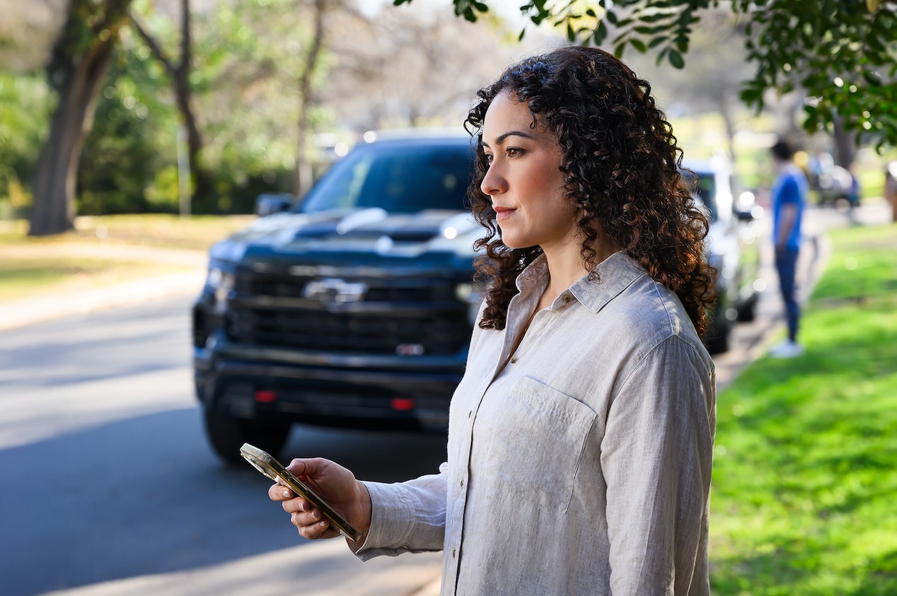 A Woman Holding a Smartphone While Standing Near a Chevy Vehicle Parked on the Street