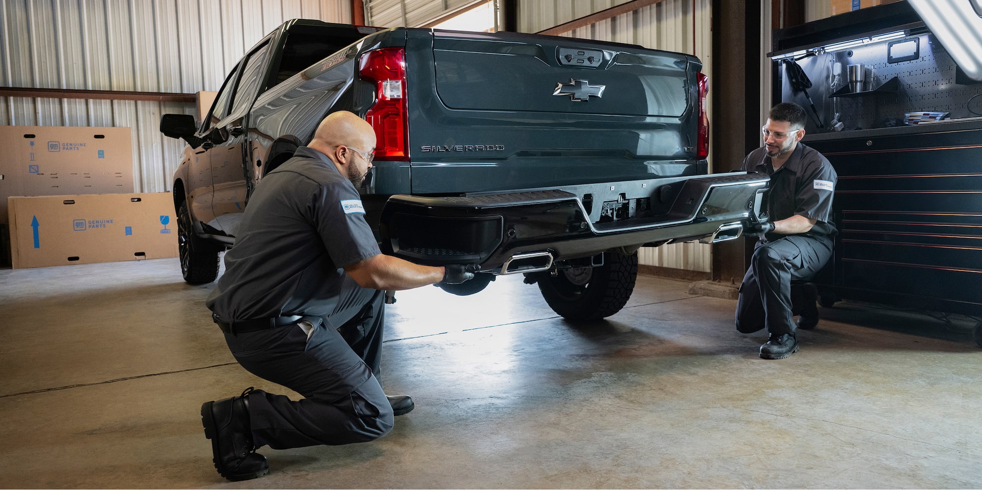 Two Auto Mechanics in a Auto Shop Install a Bumper on the Back of a Dark Gray Chevrolet Pickup Truck.