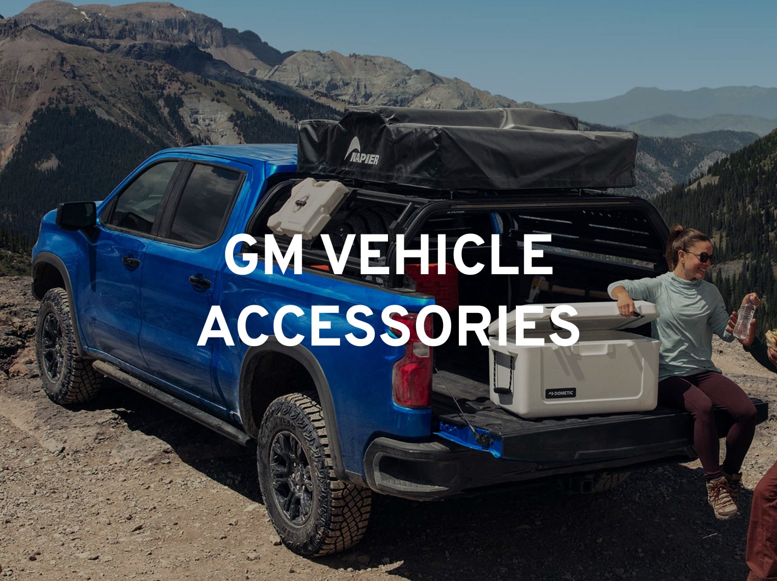 A Person Sitting In The Back Of A Blue Pickup Truck Equipped With Camping Accessories With The Text "GM Vehicle Accessories" Overlayed.
