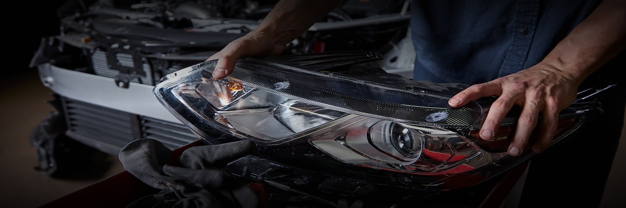 A Mechanic Holding a Headlight with a Vehicle Taken Apart Behind Him