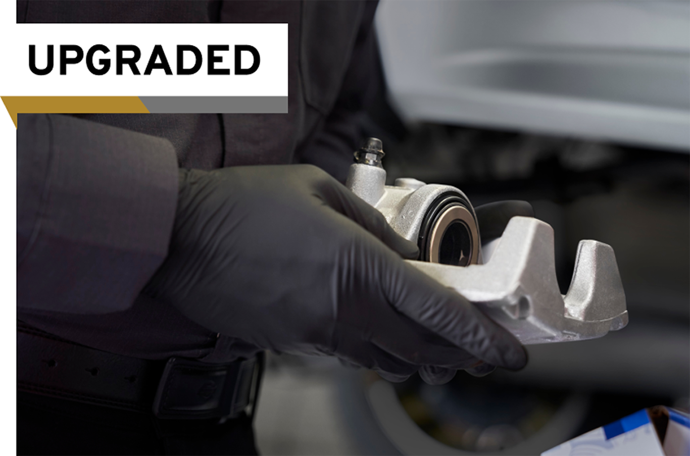 A Car Service Technician Wearing Black Gloves Holds a Disc Brake Caliper With an "Upgraded" Label on the Image.