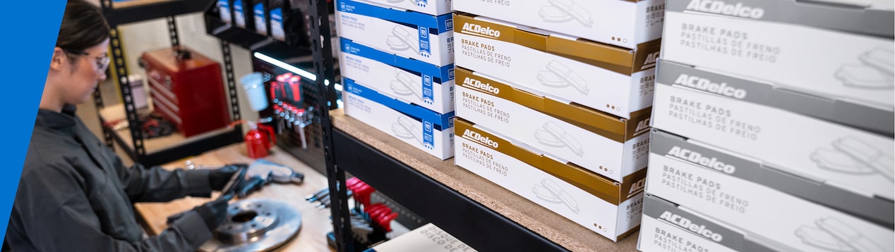 GM Technician Inspecting a Brake Rotor with Boxes of Parts Nearby on a Shelf