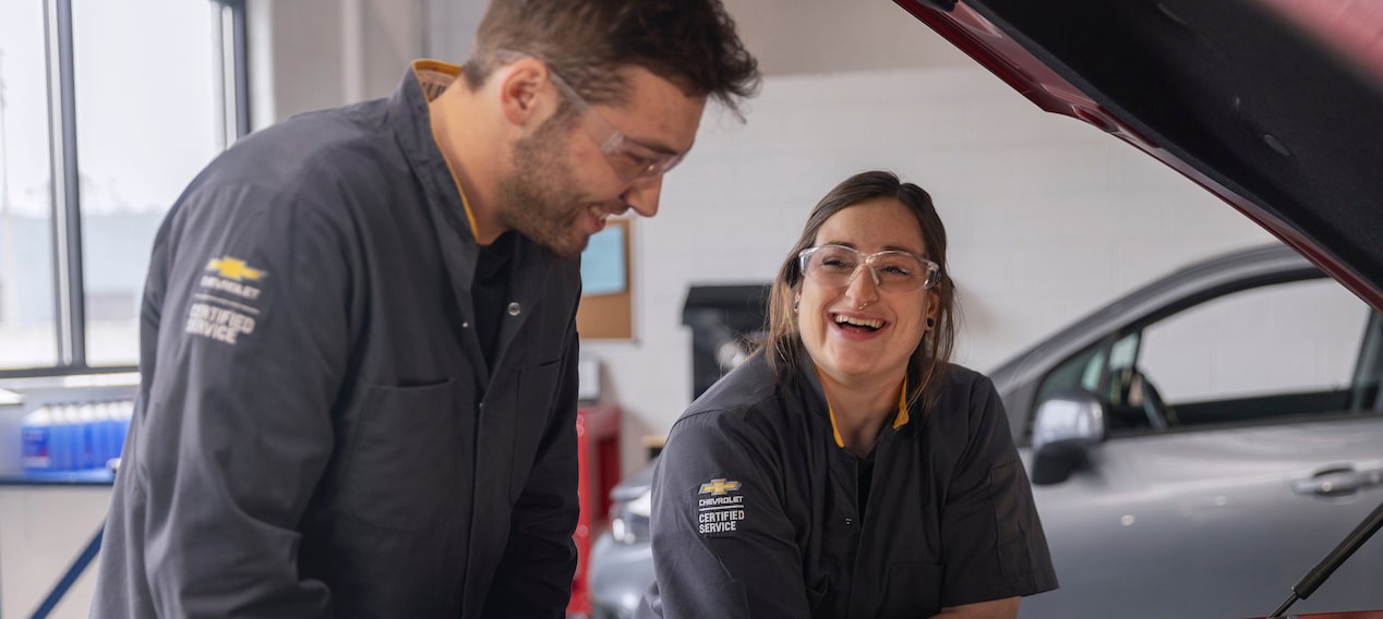 Two Smiling General Motors Certified Service Technicians in Safety Glasses Looking at a Laptop