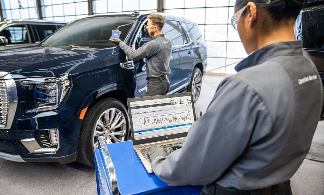 A Mechanic is Working on a Laptop While Another Mechanic is Seen Inspecting an SUV at a Distance